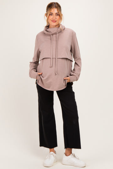 woman wearing a taupe maternity hoodie with a cowl neck and side pockets, paired with black wide-leg pants