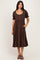 woman wearing a brown midi dress with puff sleeves, a scoop neckline, and button details, standing with one hand in her pocket