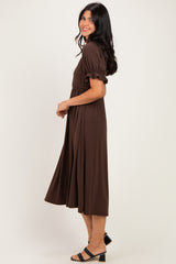 woman wearing a brown midi dress with puff sleeves and a cinched waist, standing sideways with a smile