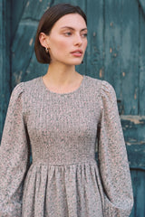 woman wearing a smocked floral print dress with long sleeves and a round neckline standing against a teal wooden background