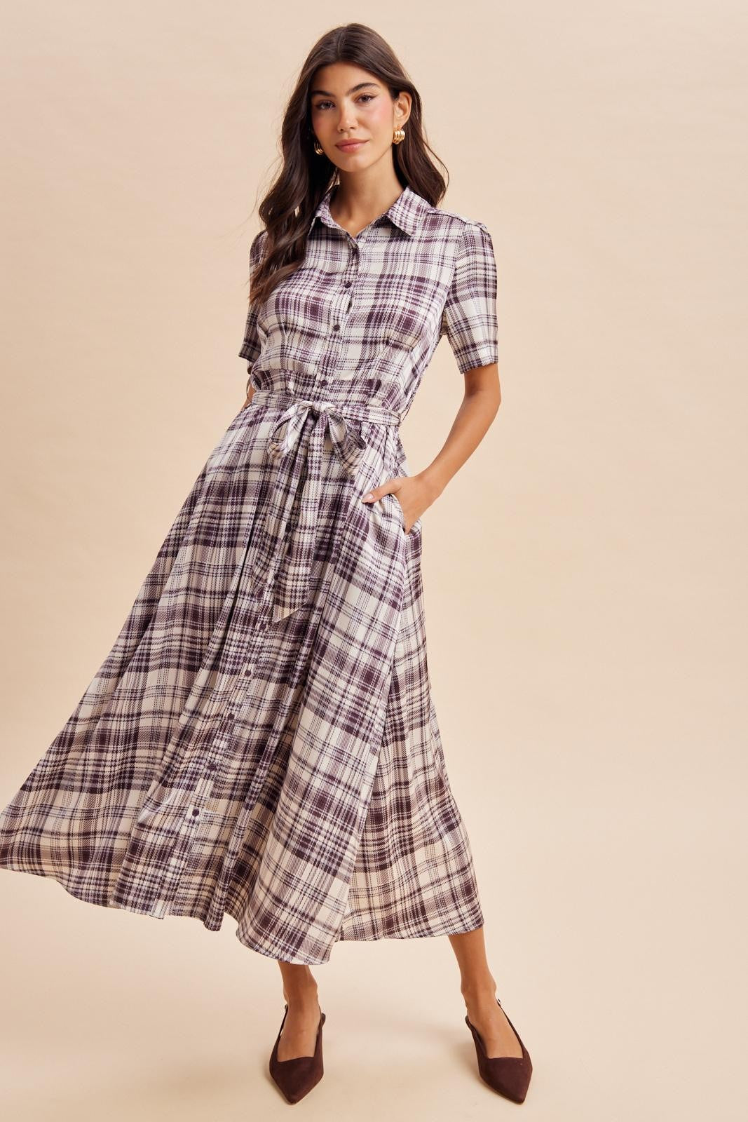 woman wearing a long plaid dress with short sleeves, button-down front, and a tied waist, standing against a beige background