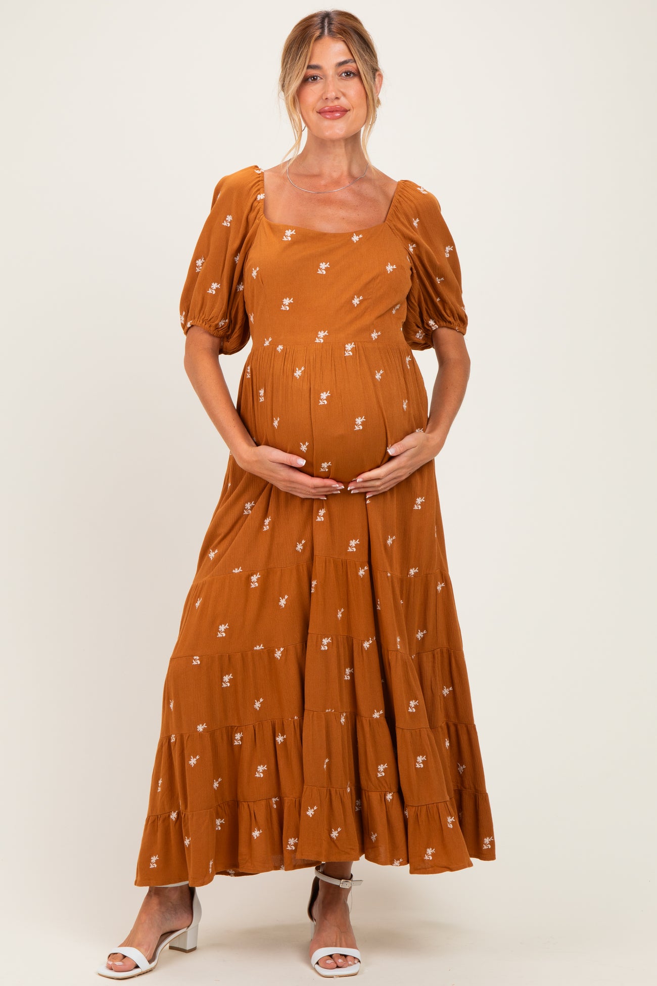 woman wearing a rust-colored maternity maxi dress with puff sleeves and floral embroidery, cradling her baby bump