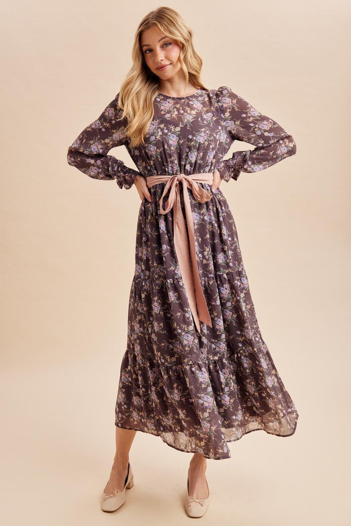woman wearing a long sleeve dark floral maxi dress with a thigh-high slit and a pink ribbon tie at the waist