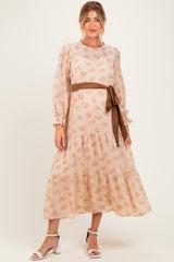 woman wearing a floral print maternity dress with long sleeves and a tied brown belt, standing in a light beige color