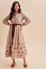 woman wearing a brown and beige patterned long sleeve dress with floral embroidery and a waist tie, posing against a neutral background