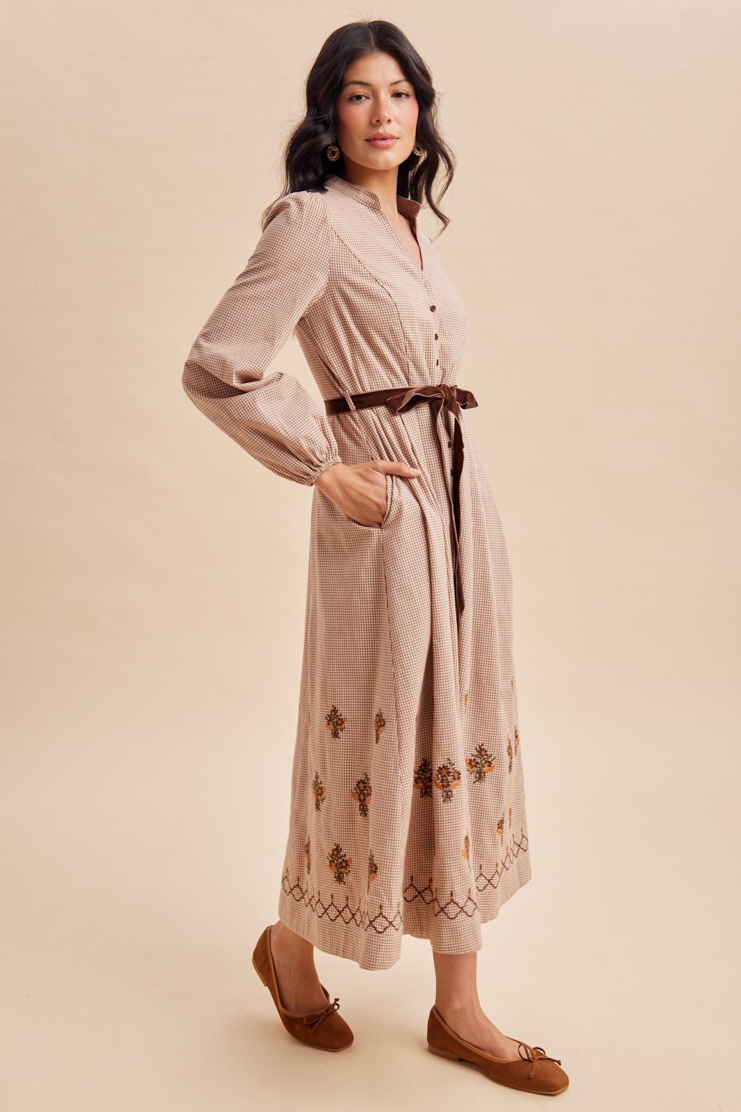 woman wearing a beige button-up midi dress with floral embroidery and a tie belt, paired with brown flats