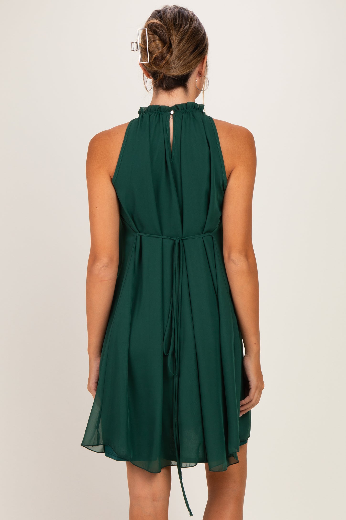 woman wearing a dark green sleeveless maternity dress with a ruffled neckline and tie-back detail