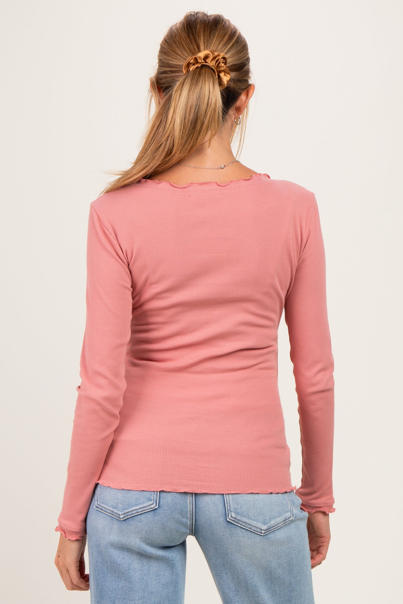 woman wearing a pink long sleeve maternity top with scalloped edges, seen from the back, paired with light blue jeans