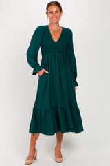 woman wearing a dark green long sleeve dress with smocked bodice and tiered skirt, posing with her hands in pockets