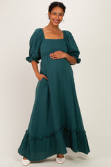 woman wearing a dark green maternity maxi dress with puff sleeves and ruffle hem, cradling her baby bump