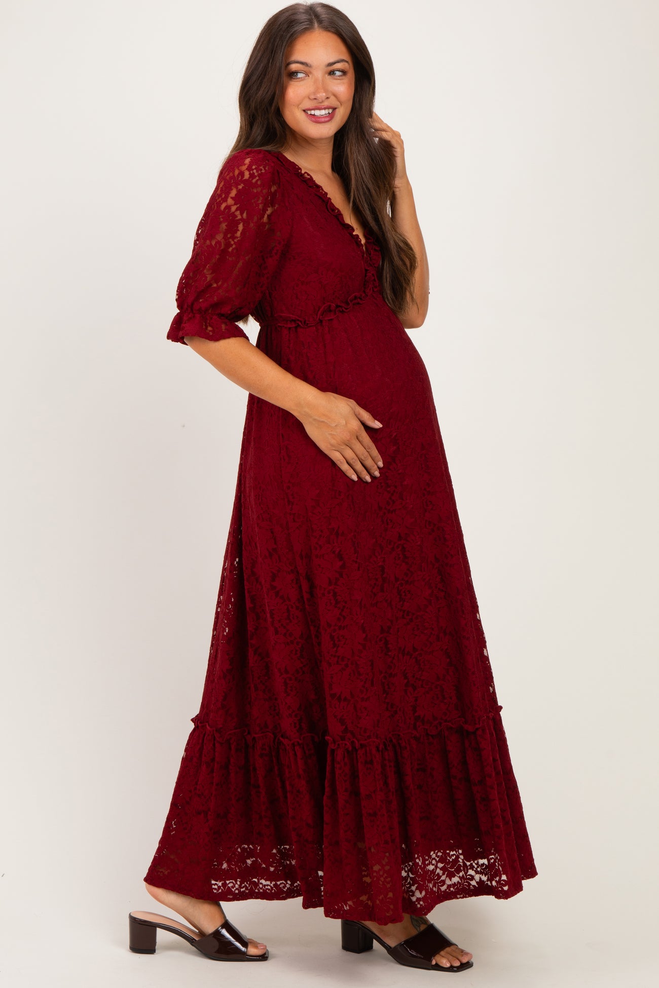 woman wearing a burgundy lace maternity dress with short sleeves and a tiered skirt, gently holding her baby bump