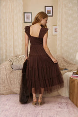 women wearing a brown tiered midi dress with frill details and a low back, standing in a cozy room