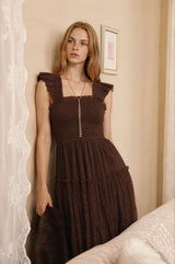 woman wearing a brown tiered dress with ruffled straps and a fitted bodice, standing by a wall with lace curtains