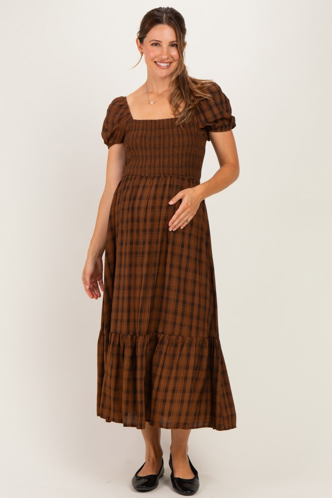woman wearing a brown plaid maternity maxi dress with puff sleeves and a tiered hem, smiling and holding her baby bump