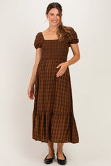 woman wearing a brown plaid maternity maxi dress with puff sleeves and a tiered hem, smiling and holding her baby bump