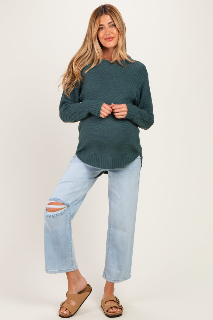 woman wearing a teal maternity sweater and distressed light blue jeans, showing her baby bump while standing casually