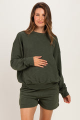 woman wearing a dark green maternity sweatshirt and matching shorts, holding her baby bump