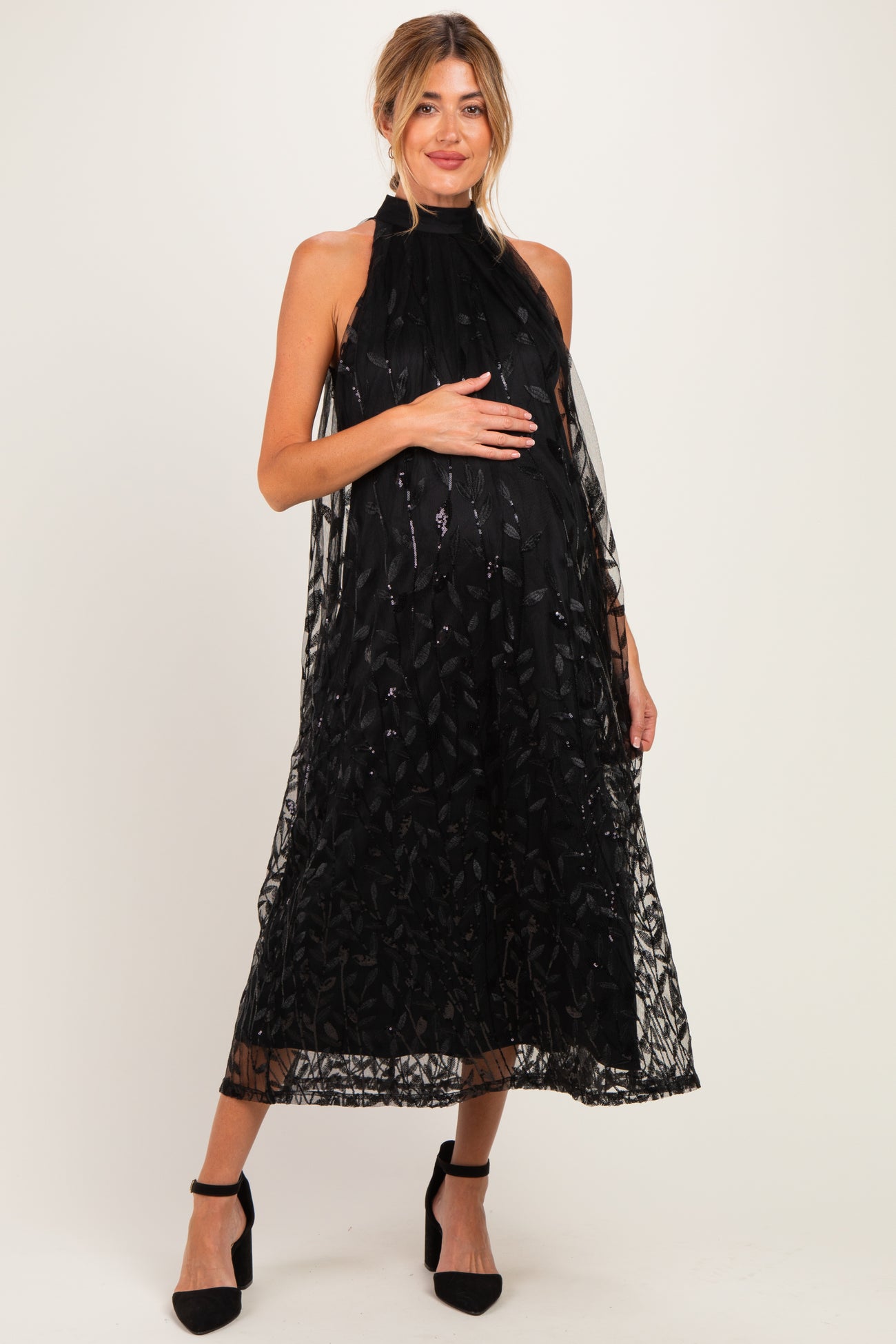 woman wearing a black maternity dress with a sheer overlay and floral details, showcasing her baby bump