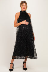 woman wearing a black maternity dress with a sheer overlay and floral details, showcasing her baby bump