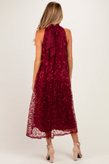 woman wearing a burgundy maternity dress with floral embroidery and a tie-back detail, showing a flowing length and a sleeveless design