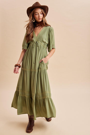 woman wearing a green maxi dress with short puff sleeves, a v-neck, and a tied waist detail, paired with a brown hat