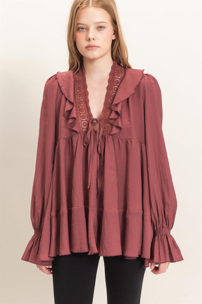 woman wearing a burgundy long sleeve top with ruffle details and a tie neckline, paired with black pants