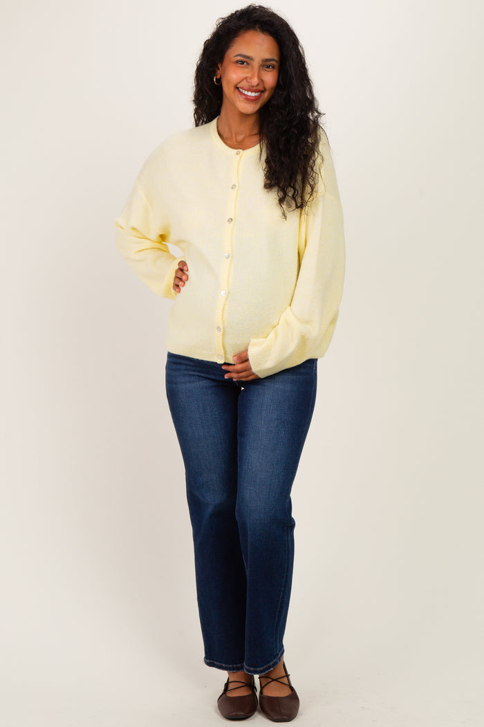 woman wearing a yellow maternity cardigan and blue jeans, gently holding her baby bump while smiling