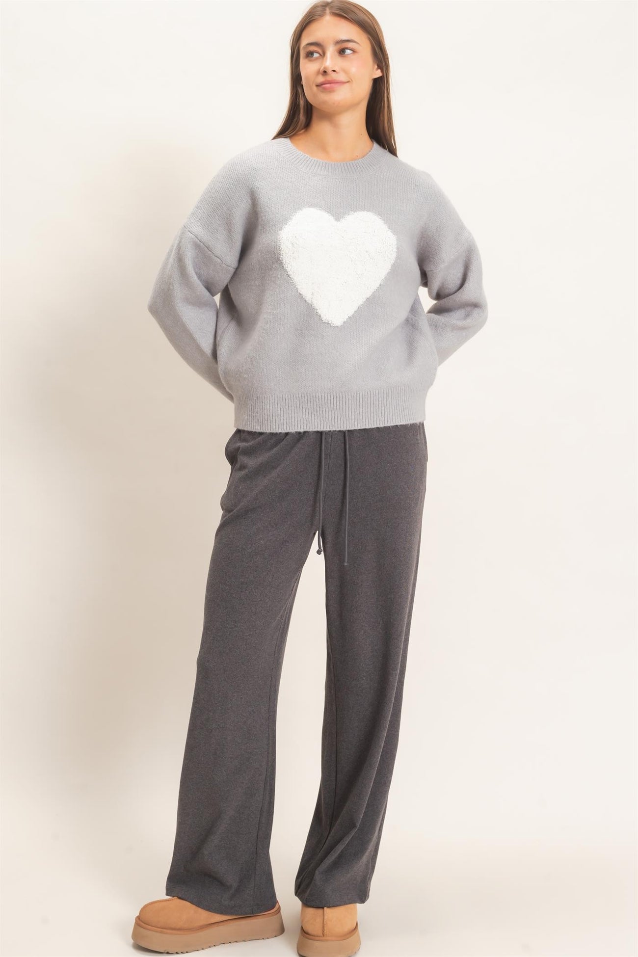 woman wearing a grey sweater with a white heart graphic and grey wide-leg pants, standing with hands on hips
