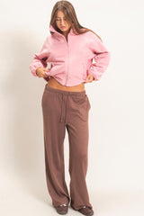 woman wearing a pink cropped fleece jacket and brown wide-leg pants, looking down while adjusting her outfit