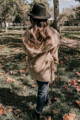woman wearing a tan fuzzy hooded jacket, black hat, and dark jeans, standing in a park surrounded by autumn leaves