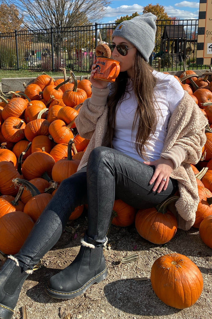 woman sitting among pumpkins wearing a white t-shirt, black jeans, and a cozy beige cardigan, drinking from a mug