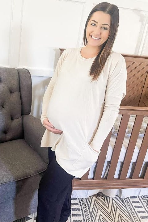 woman wearing a beige long sleeve maternity top and black pants, smiling while holding her baby bump.