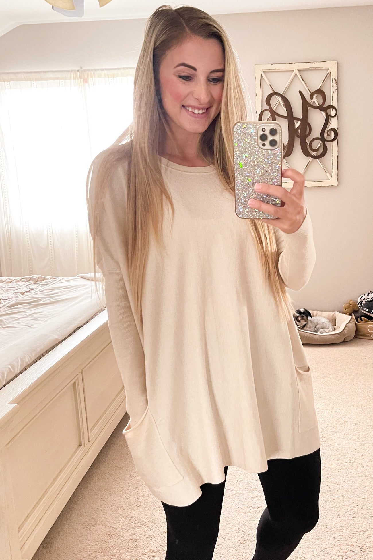 woman wearing a beige oversized sweater with side pockets and black leggings, smiling while taking a mirror selfie