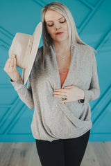 woman wearing a light grey maternity wrap top and black leggings, holding a beige hat and showing her baby bump
