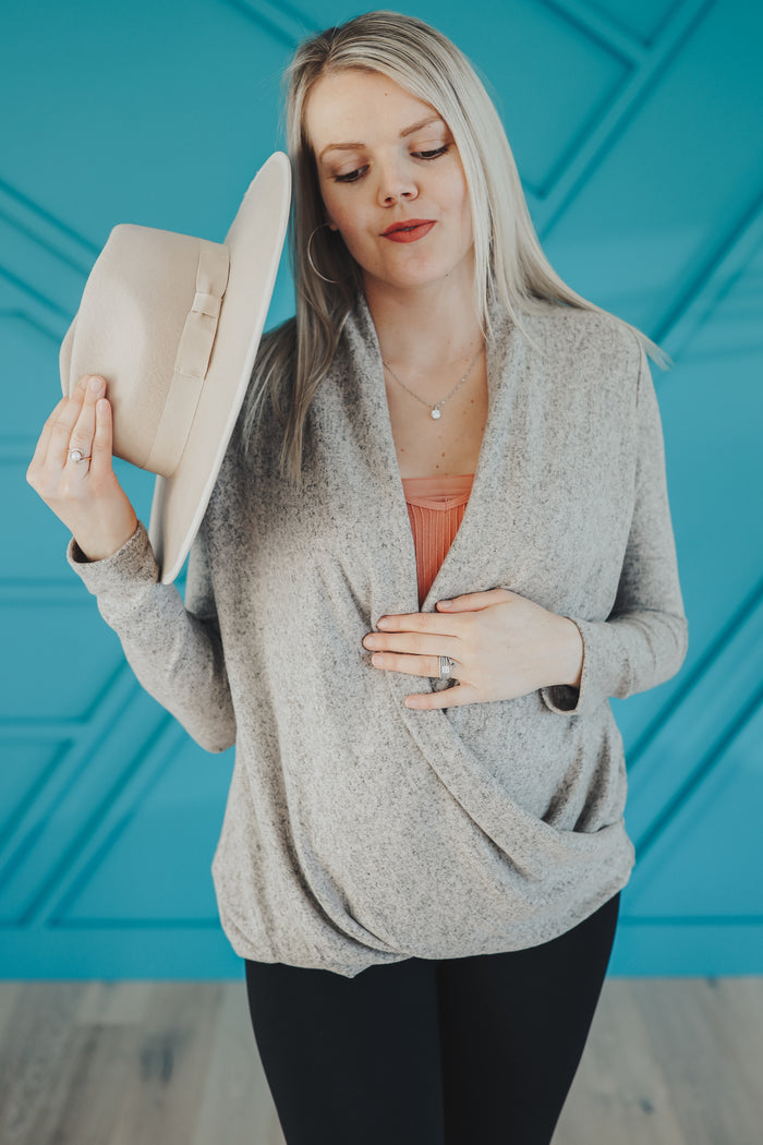 woman wearing a light grey maternity wrap top and black leggings, holding a beige hat and showing her baby bump