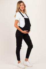 woman wearing a black maternity overall with ripped details and a white short sleeve top, holding her baby bump.
