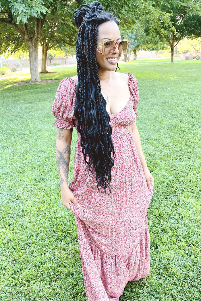 woman with long black braids wearing a pink floral maxi dress and oversized sunglasses in a park