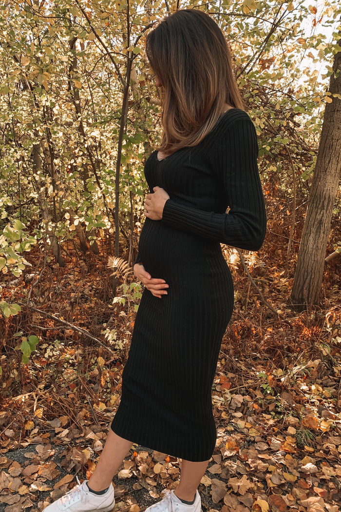 woman wearing a black ribbed maternity dress and holding her baby bump, standing in a forest with autumn leaves
