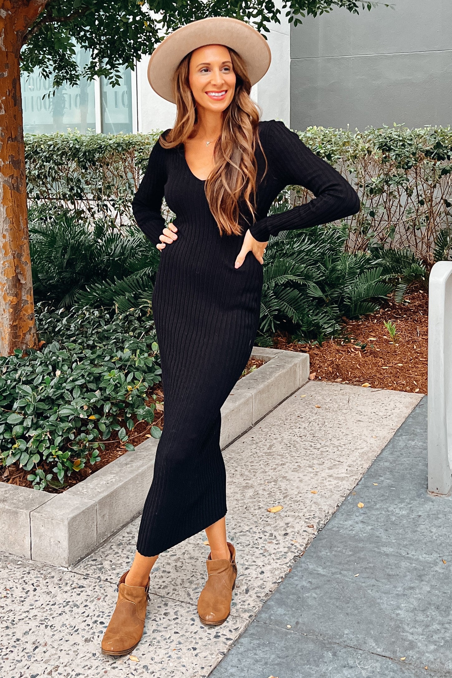 Woman in a black ribbed midi dress, tan wide-brimmed hat, and brown ankle boots posing outdoors