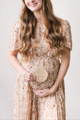 woman wearing a floral maternity dress holding a sign that says "can't wait to meet you" with her baby bump visible