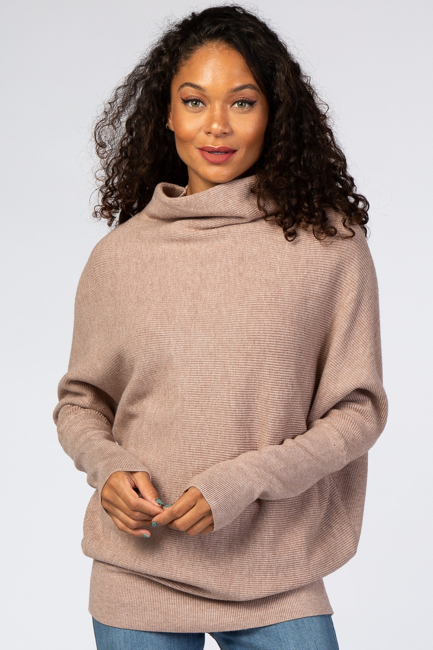 woman wearing a light brown oversized sweater with a cowl neck and long sleeves, paired with blue jeans