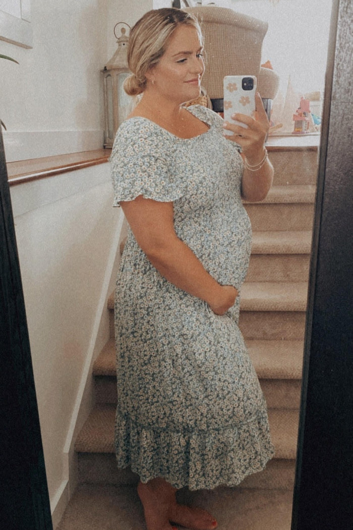 woman wearing a light blue floral maternity dress, gently holding her baby bump while standing by a staircase