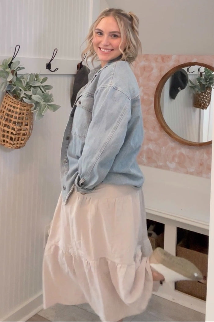 woman wearing a light denim oversized jacket and a flowy beige tiered skirt, smiling and standing in a bright room