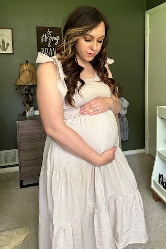 woman wearing a light pink maternity dress with ruffled sleeves, cradling her baby bump while standing indoors