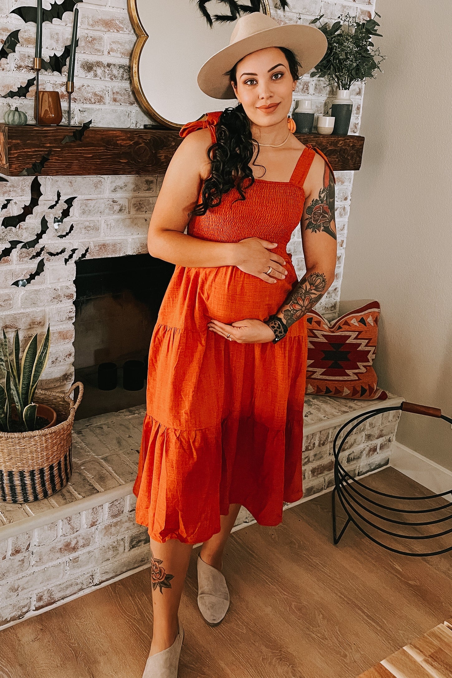 woman wearing an orange tiered maternity dress and holding her baby bump, accessorized with a wide-brimmed hat