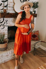 woman wearing an orange tiered maternity dress and holding her baby bump, accessorized with a wide-brimmed hat