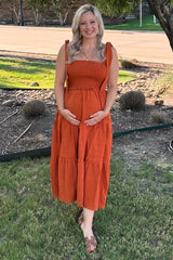 woman wearing an orange maternity dress with a smocked bodice and tiered skirt, cradling her baby bump outdoors