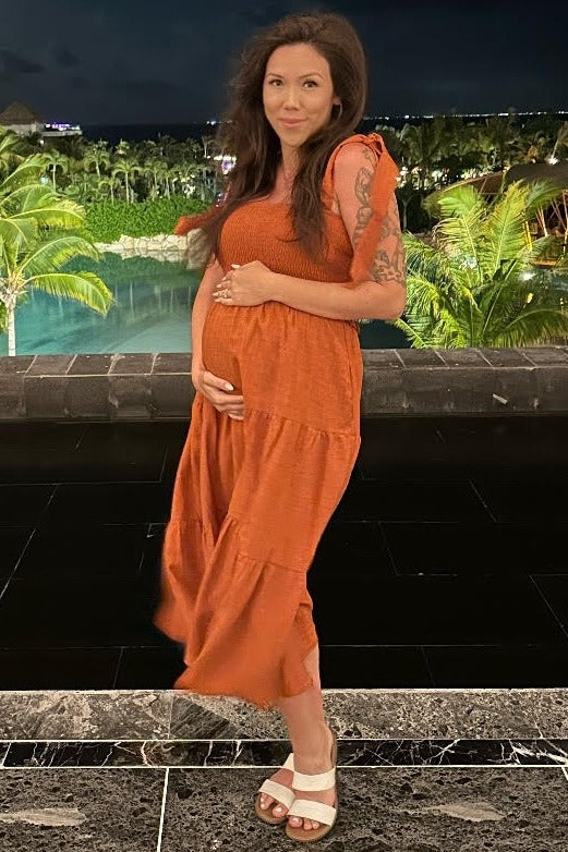 woman wearing an orange maternity maxi dress and holding her baby bump in an outdoor setting with greenery in the background