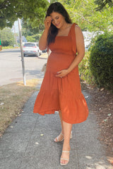 woman wearing a rust-colored maternity dress with a tiered skirt and holding her baby bump while smiling outdoors