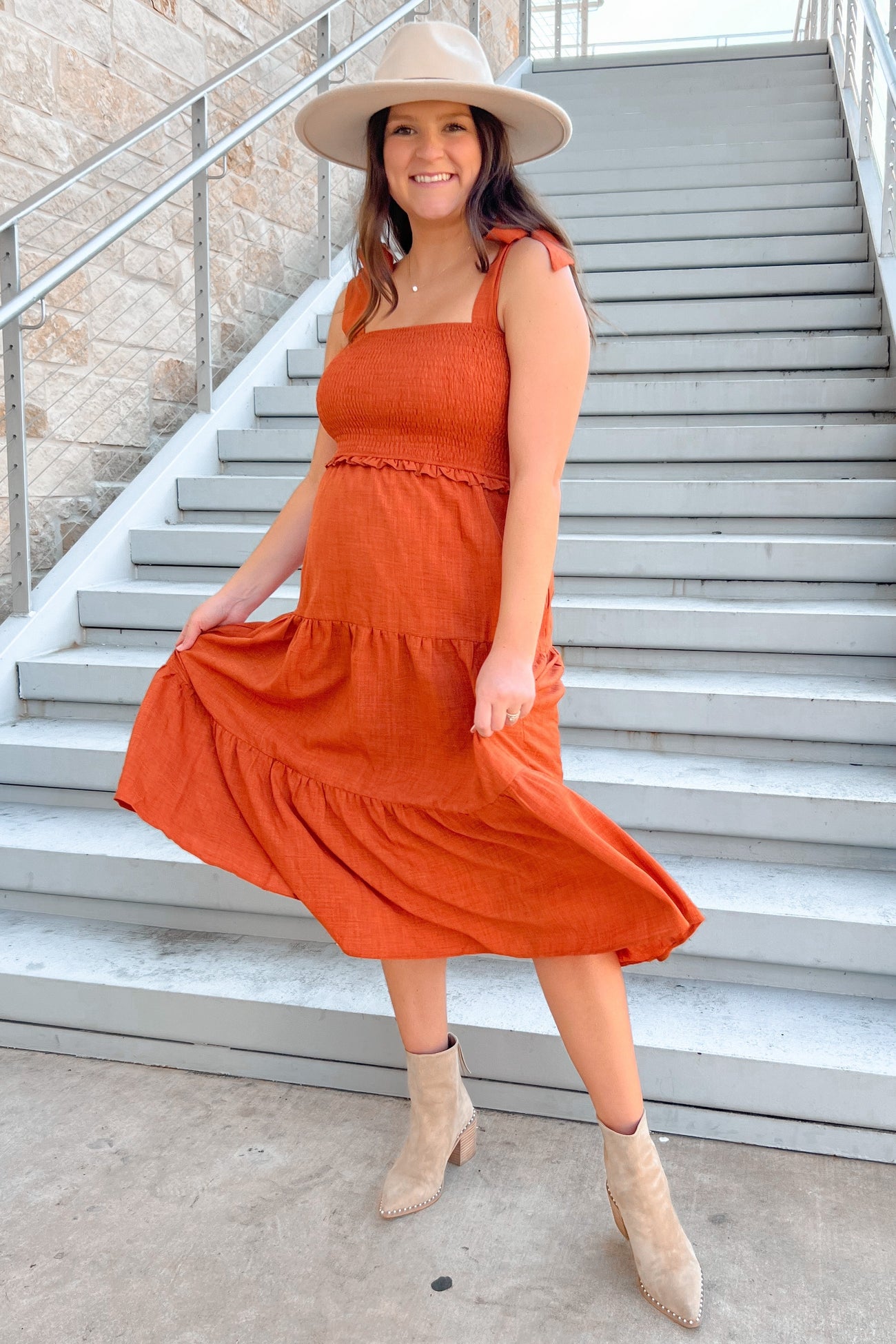 woman wearing an orange maternity dress with a smocked bodice and tiered skirt, holding her baby bump, on outdoor stairs